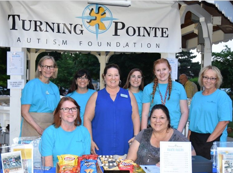 Eight people in blue shirts stand under a Turning Pointe Autism Foundation banner at an outdoor event booth.
