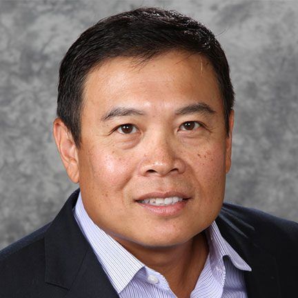 A smiling person wearing a dark blazer and a light blue patterned dress shirt against a grey textured background.