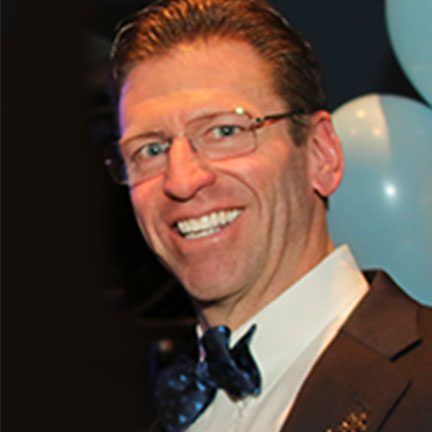 A person smiling while wearing glasses, a light shirt, and a dark blue patterned bow tie, with balloons in the background.