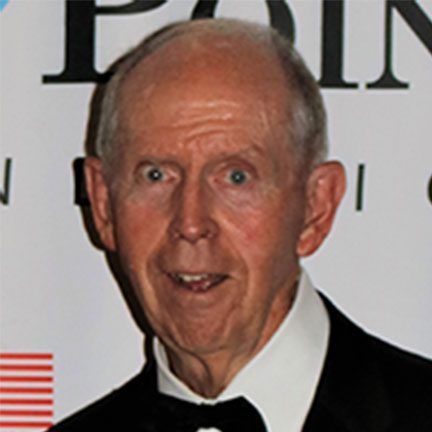 A person in a black tuxedo and white shirt posing in front of a white backdrop with partial text.