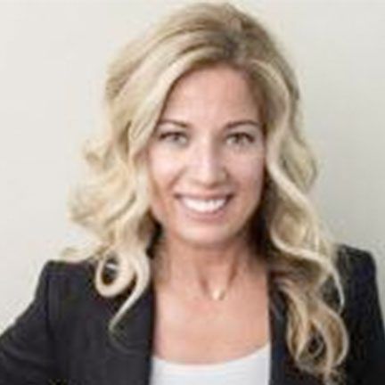 A person with shoulder-length blonde wavy hair, wearing a dark blazer over a white shirt, smiling against a plain wall.