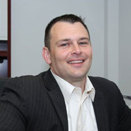 A person with short hair smiles, wearing a white collared shirt and a dark pinstriped blazer in an office setting.