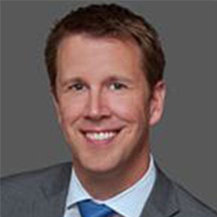 A headshot of a person smiling, wearing a suit and a blue tie, against a solid gray background.