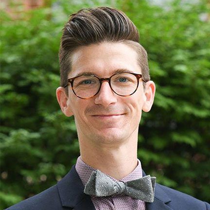 A person wearing glasses and a grey bowtie, smiling against a background of green foliage.