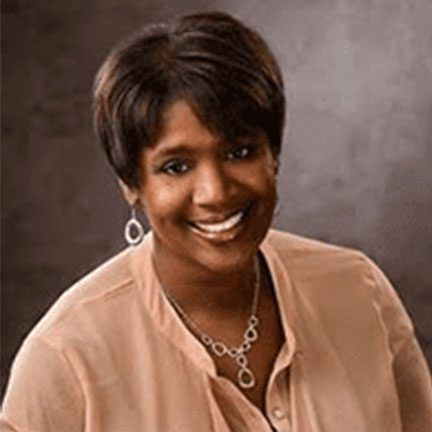 A smiling person with short dark hair, wearing a light peach-colored top and a silver necklace, against a gray background.