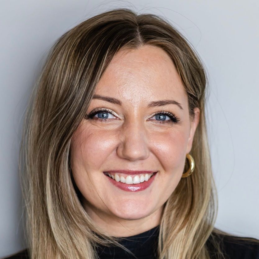 Headshot of a smiling person with shoulder-length blonde hair, wearing a black top and gold hoop earrings.