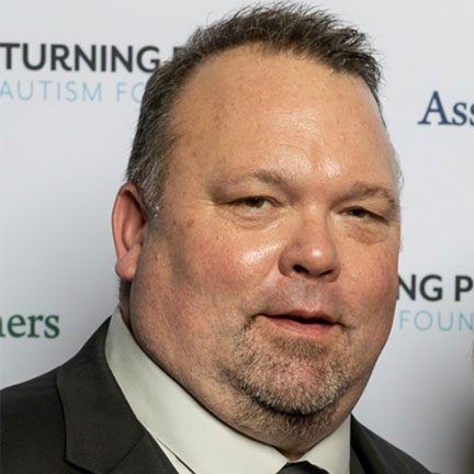 A man with short hair and a goatee wearing a suit and light shirt, standing in front of a branded event backdrop.