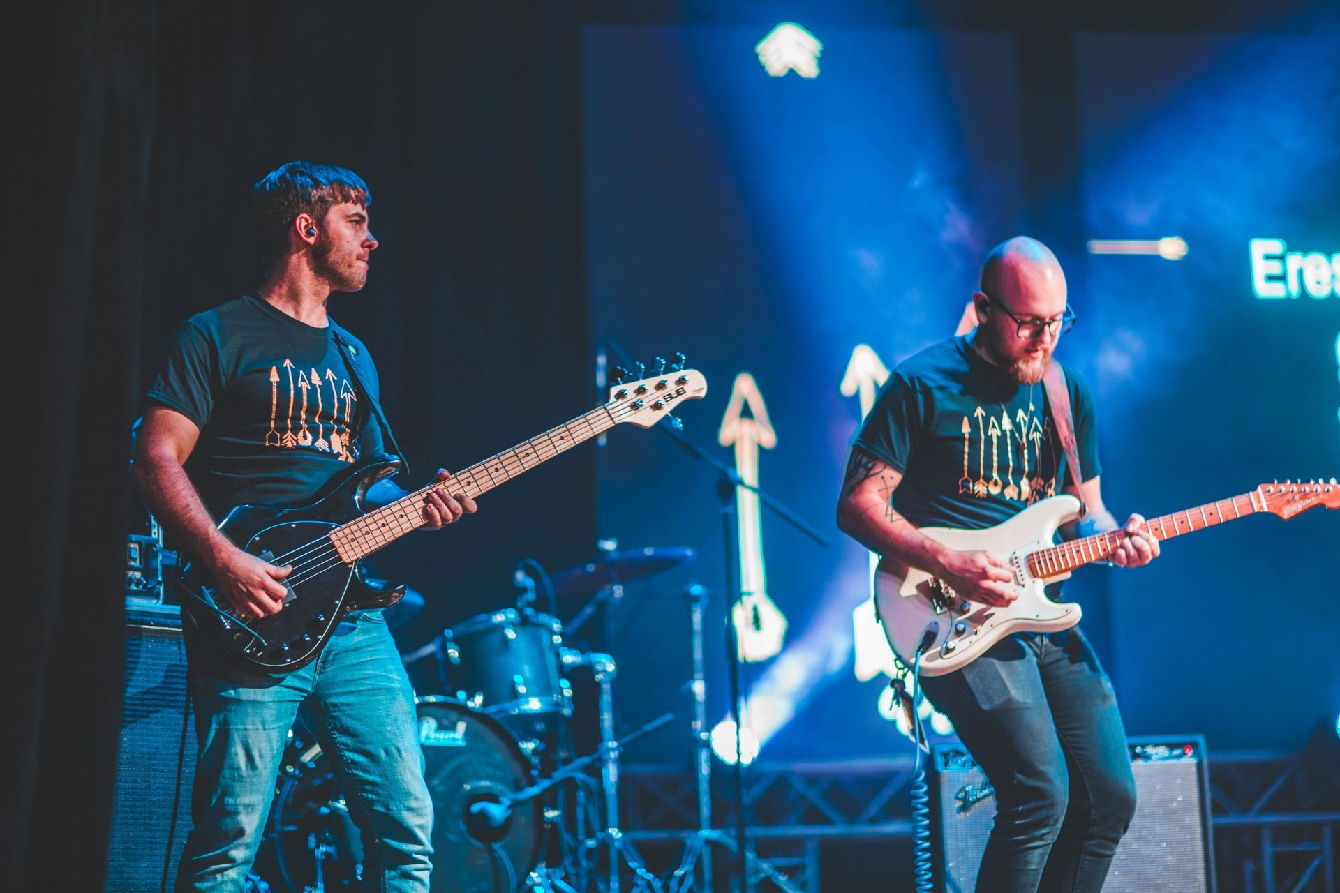 Two guitarists perform on a dimly lit stage with blue spotlights and a screen showing text in the background.