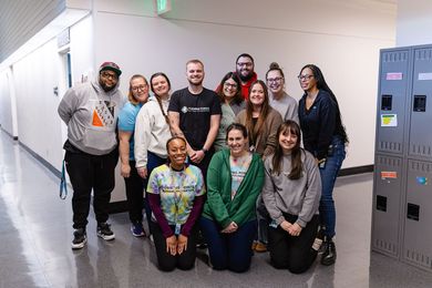 A group of fourteen people posing for a photo in a brightly lit hallway next to a row of gray metal lockers.