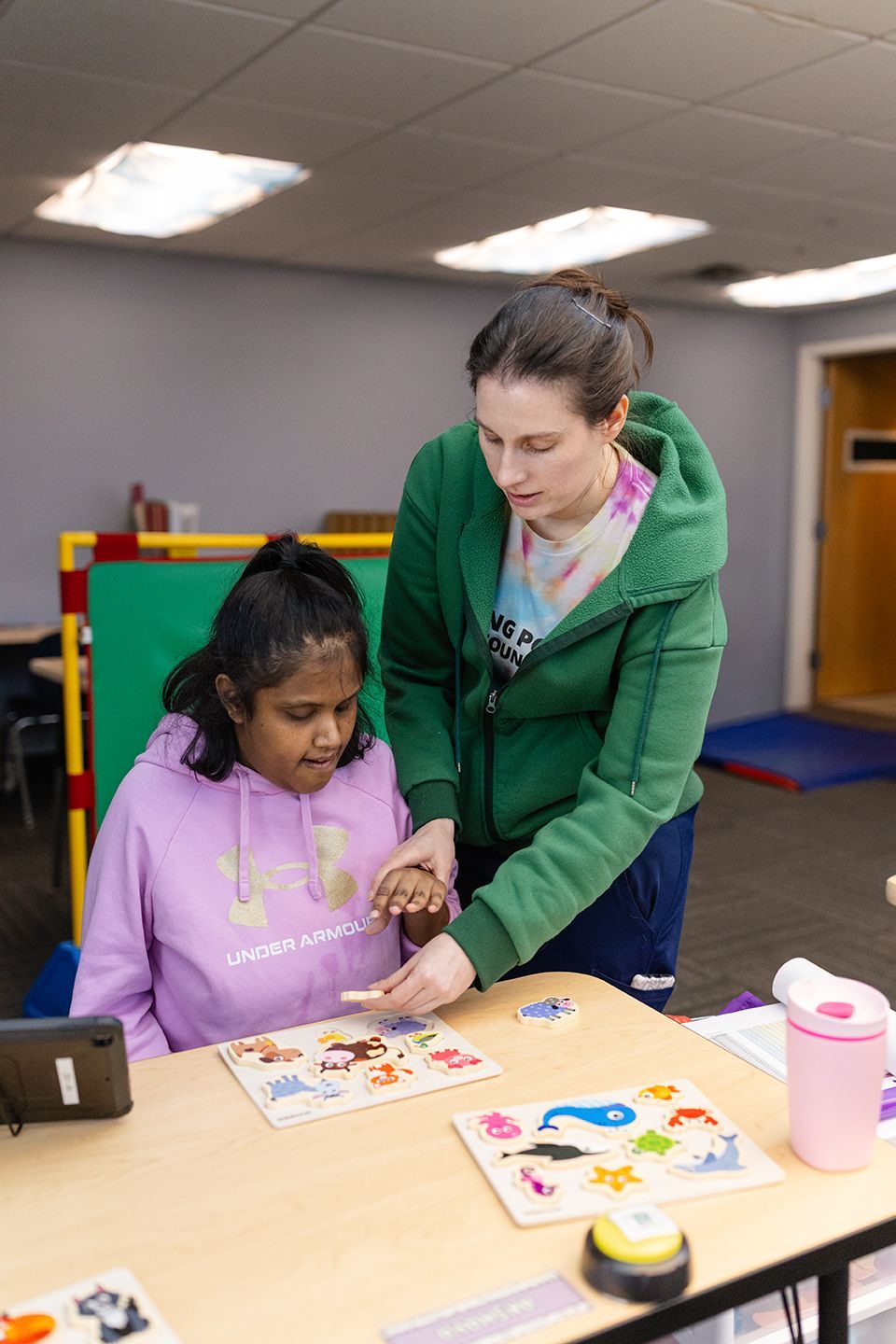 An instructor assists a student in a classroom setting as they work together on wooden puzzles on a table.