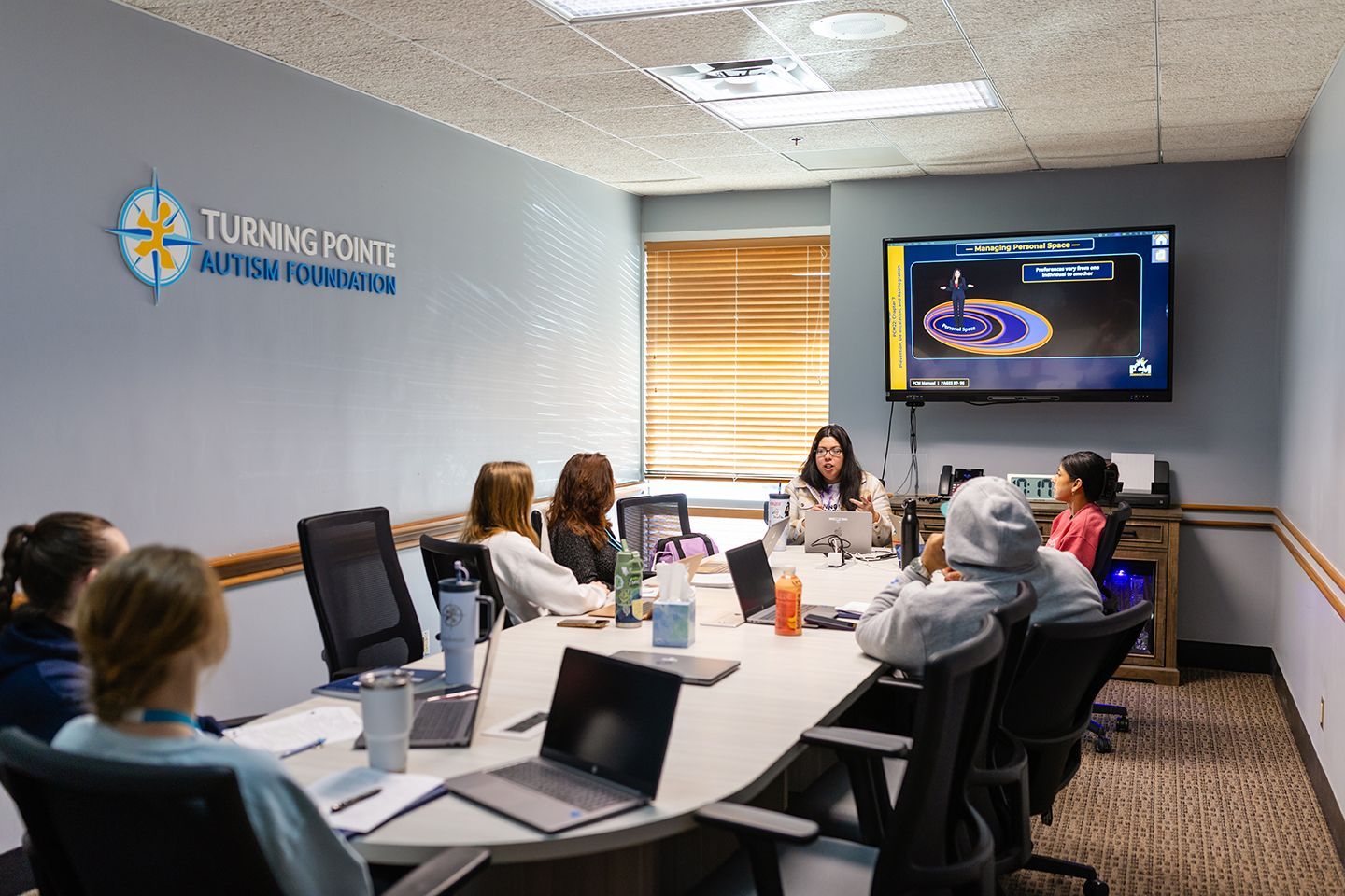 A group of people sit around a conference table in an office, facing a presenter and a screen displaying a presentation.
