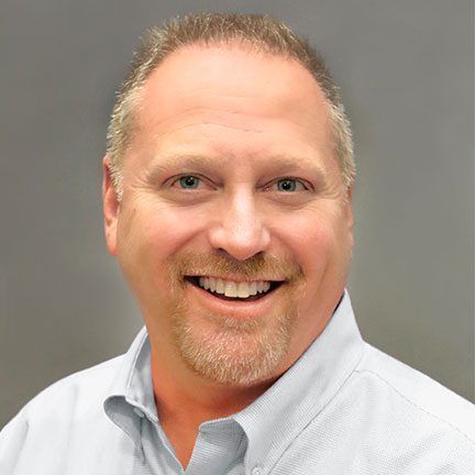 Smiling person with short hair and a goatee, wearing a light blue button-down shirt against a grey background.