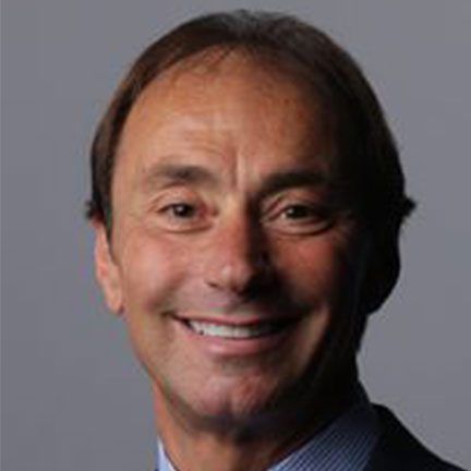 Headshot of a smiling person wearing a blue shirt and dark blazer against a plain gray background.