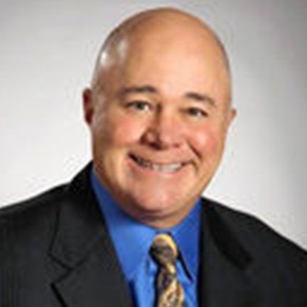 A bald person wearing a dark blue suit jacket and light blue dress shirt, smiling against a plain gray background.