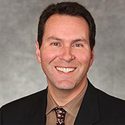 A man in a black blazer and brown collared shirt with a patterned tie, smiling against a mottled gray background.