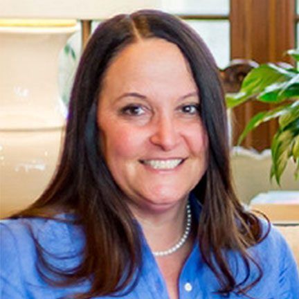 A smiling person with long dark hair, wearing a blue collared shirt and a pearl necklace, indoors with plants.
