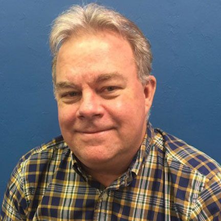 A person with light hair, wearing a blue and yellow plaid shirt, smiles gently against a solid blue background.