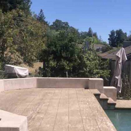 A patio with a swimming pool in the background and trees in the background.