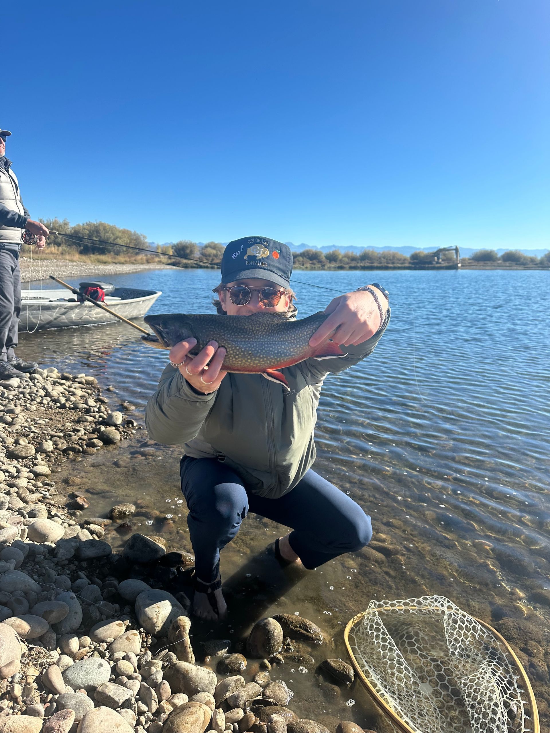 A man is kneeling on the shore of a lake holding a fish.