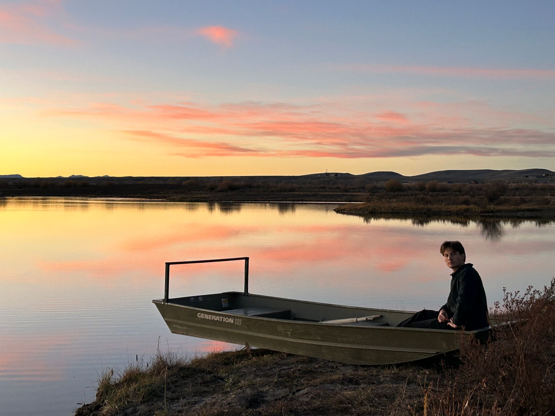 A man is sitting in a boat on the shore of a lake at sunset.