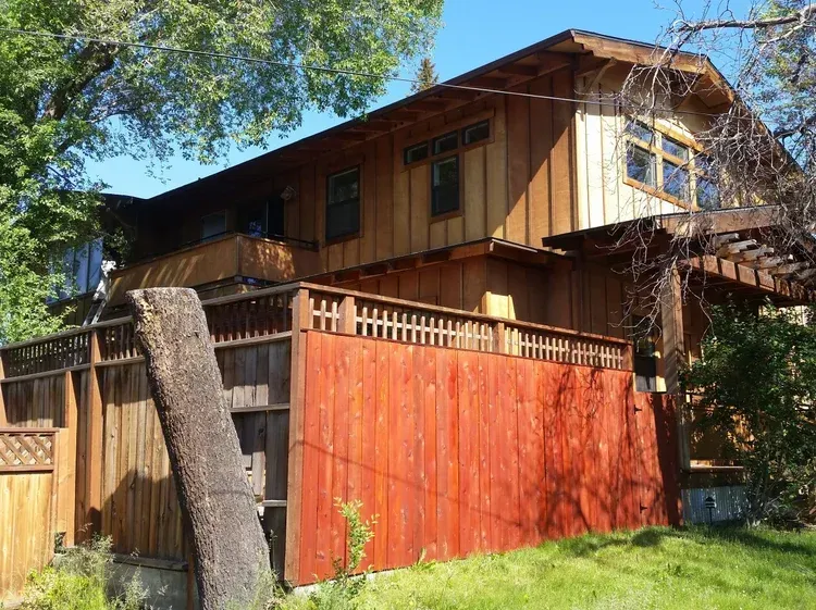 Two-story wooden house with a red fence in front and a pergola on the side, surrounded by greenery.