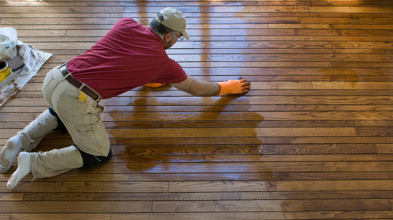 Person applying finish to hardwood floor, kneeling, wearing work clothes and gloves.