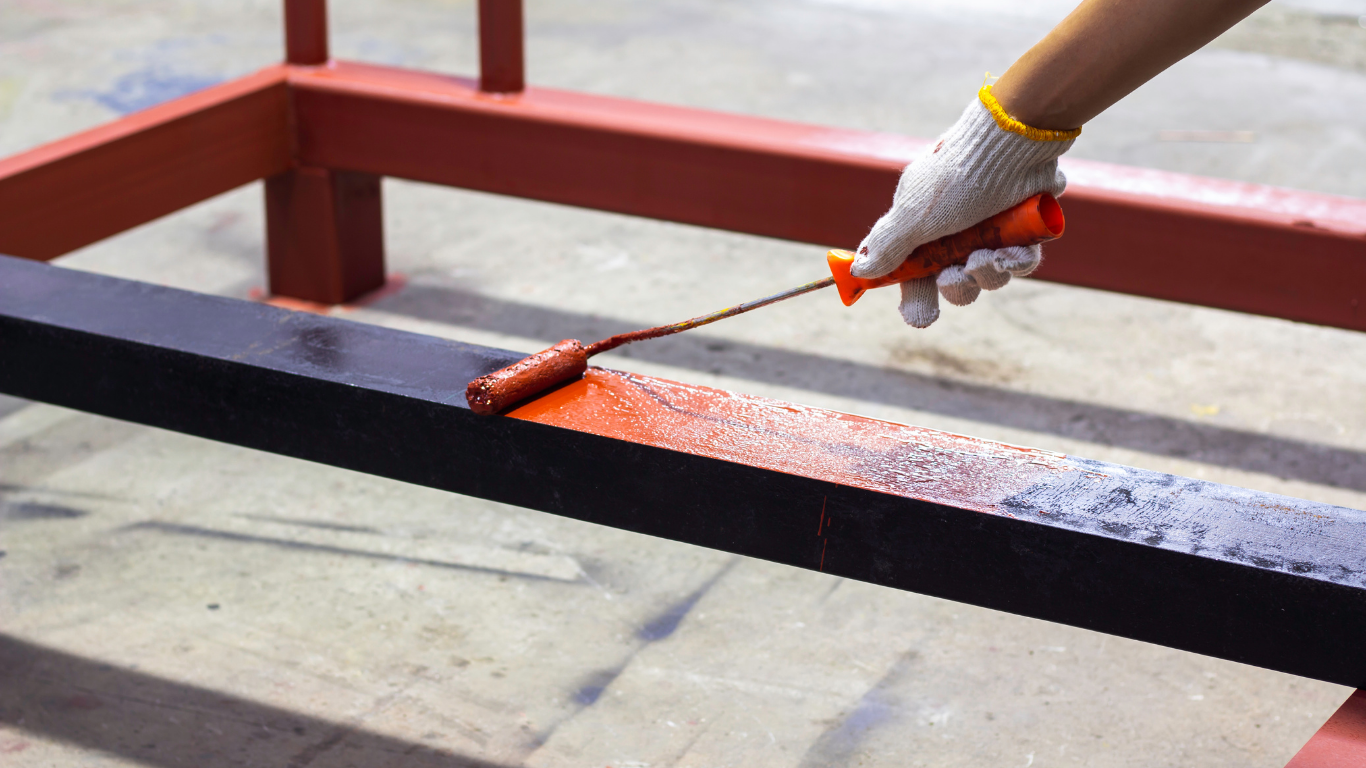 Person in glove painting a black metal beam with an orange paint roller.