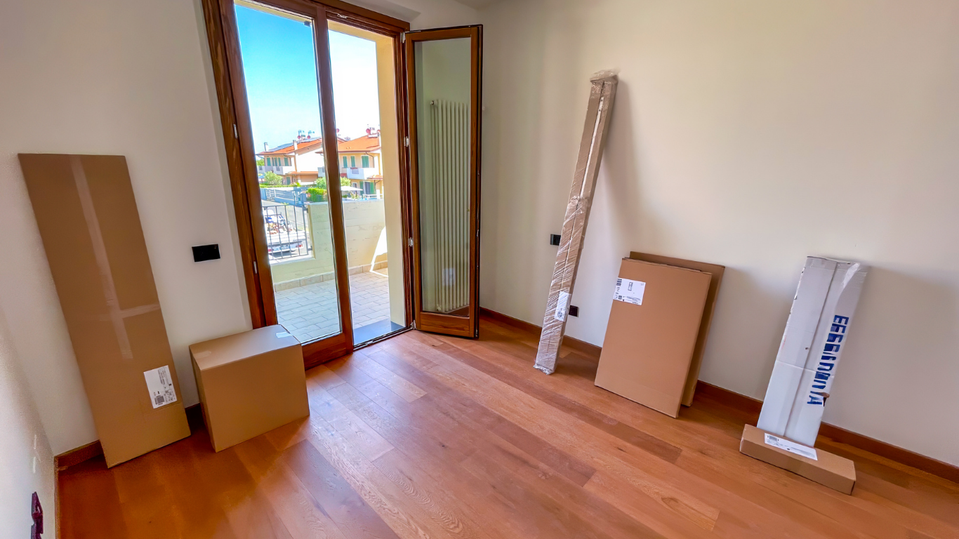 Empty room with wooden floor and balcony. Cardboard boxes and items are against the walls.