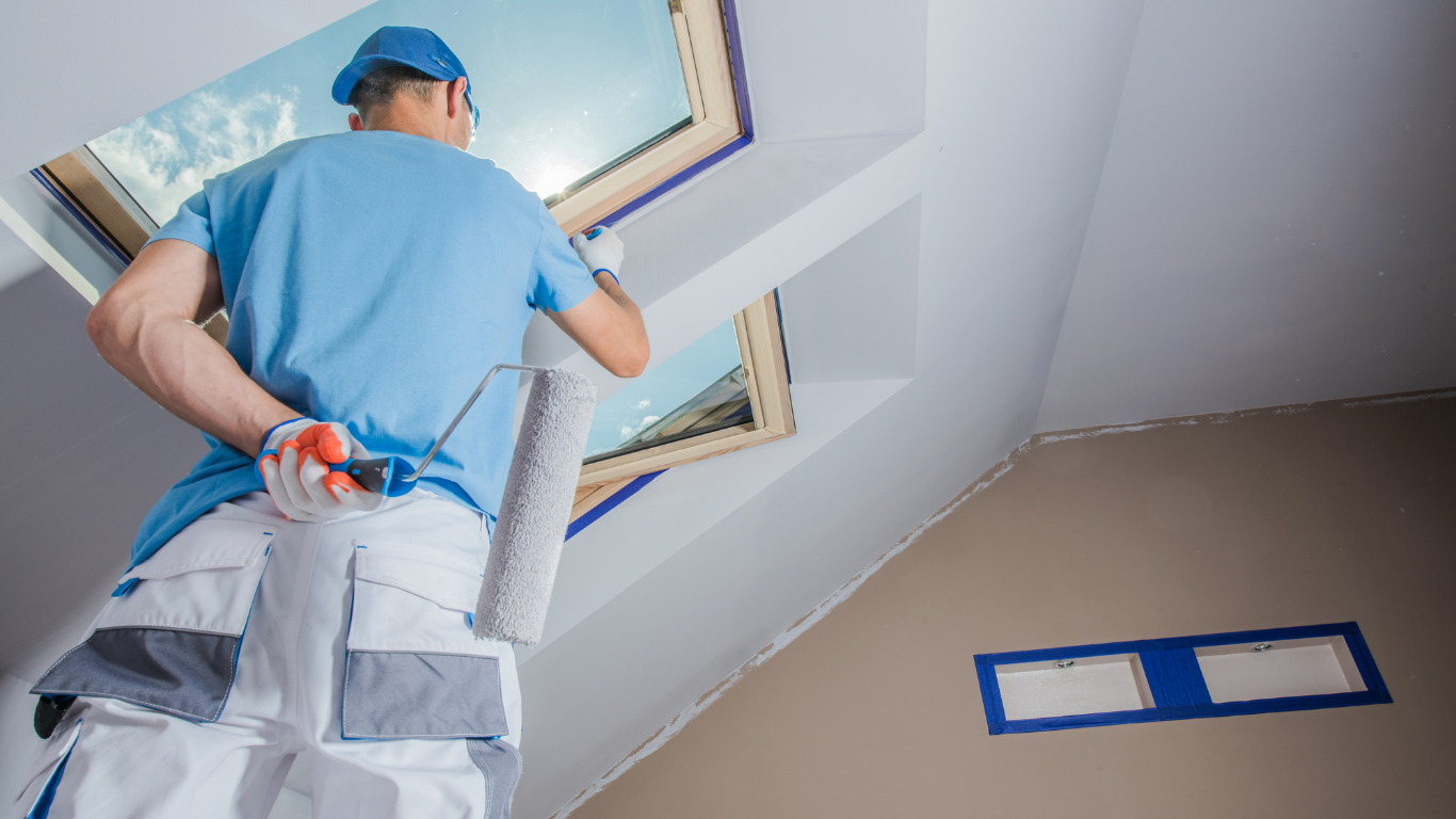 Painter on a ladder, rolling white paint around a skylight. Interior setting, blue and white colors.