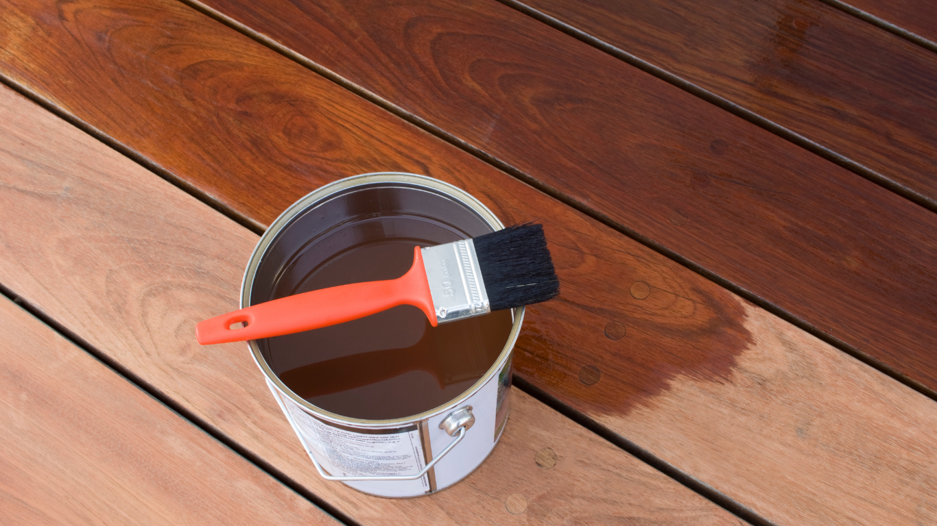 Wood stain being applied to a wooden deck with an orange-handled brush resting on a paint can.
