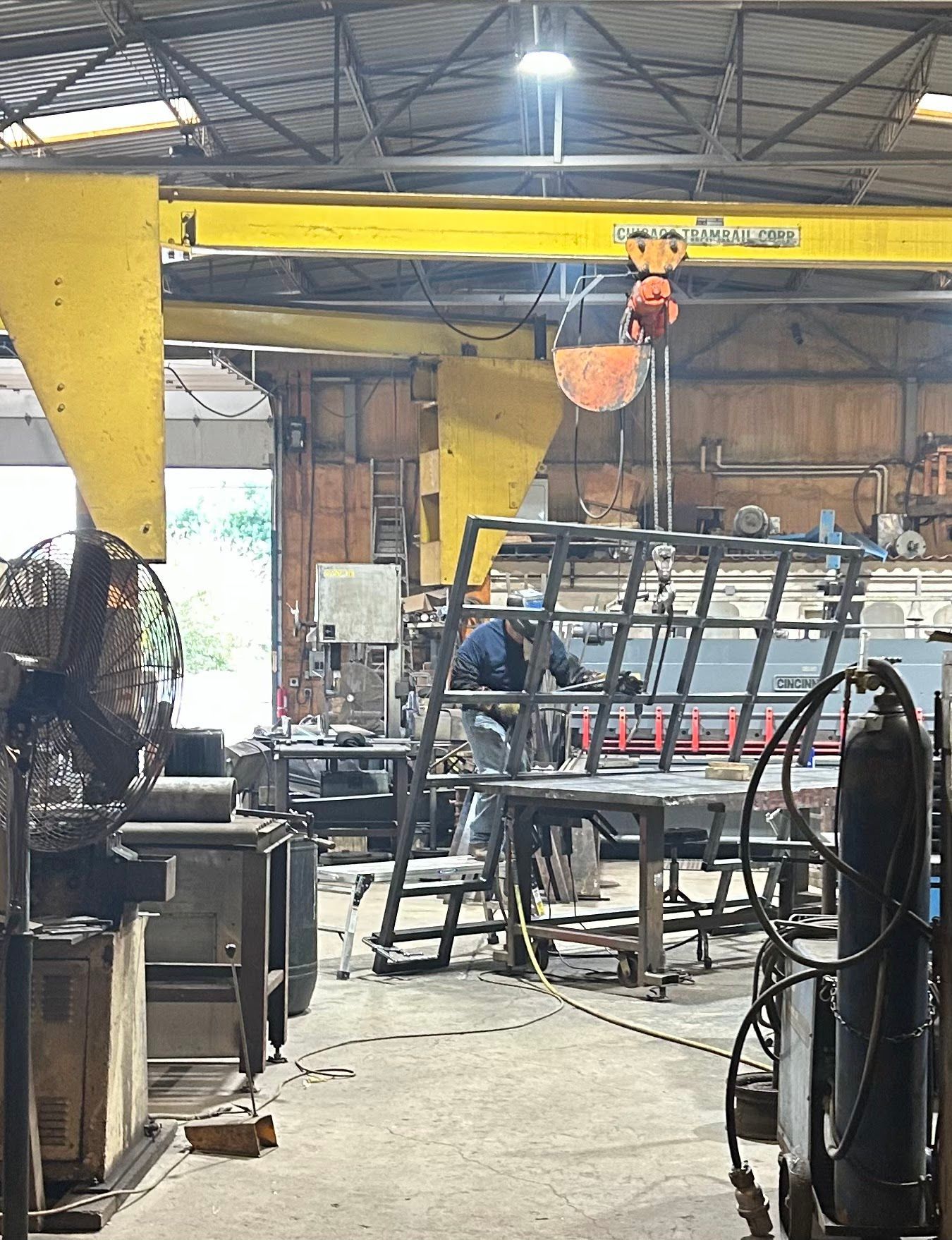 A person welding metal in a workshop under a yellow overhead crane.