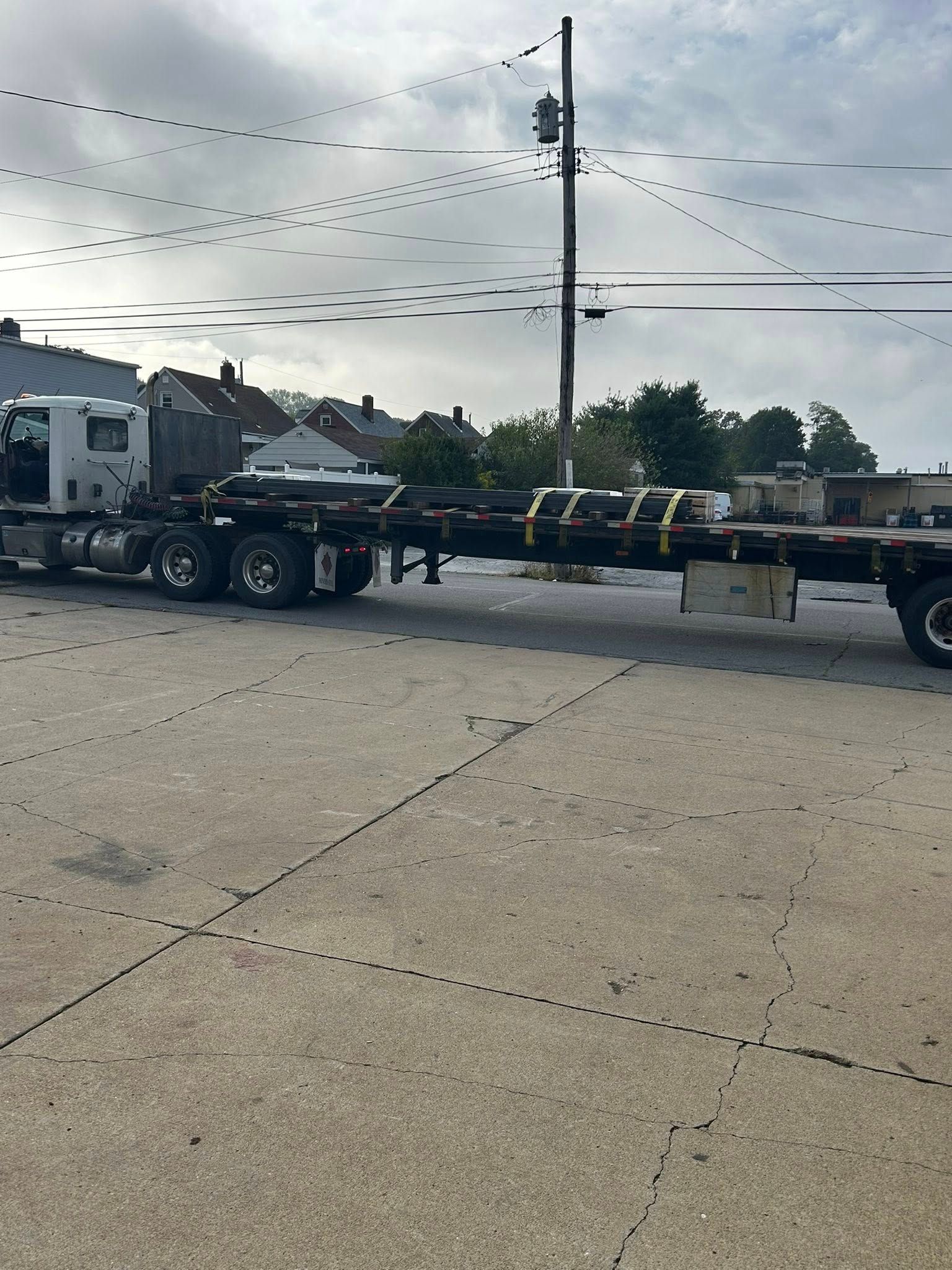 Semi-truck flatbed parked in a paved lot; power lines above; buildings in the background.