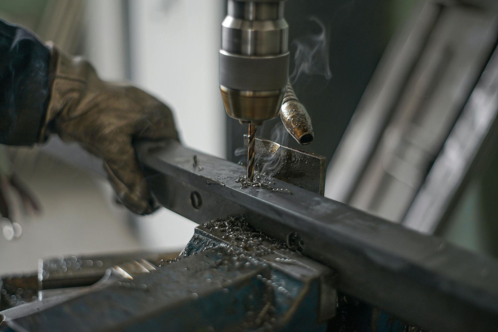 Person in work gloves drilling a metal bar with a drill press, creating shavings.