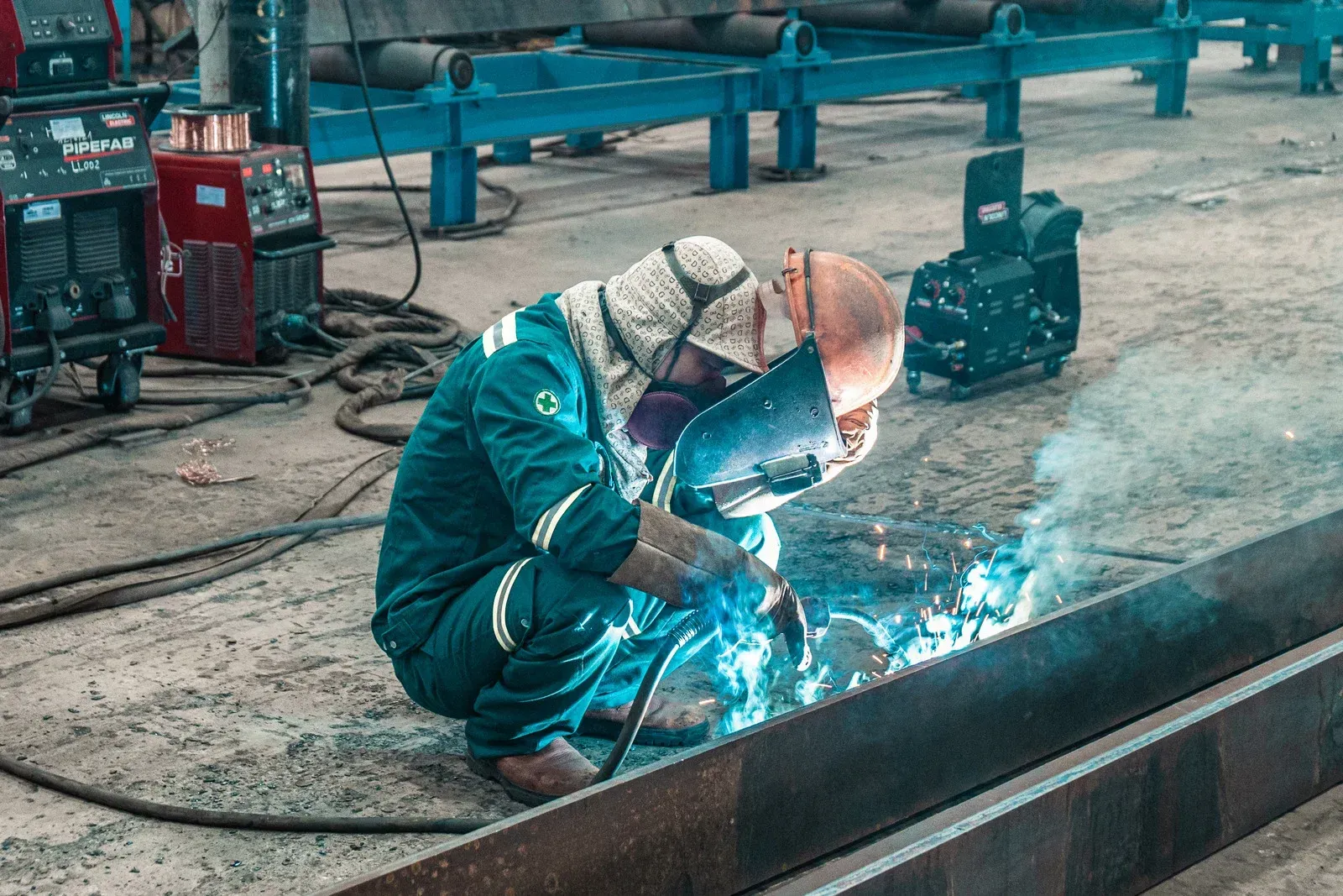 Welder in blue coveralls, kneeling, welding metal with sparks, industrial setting.