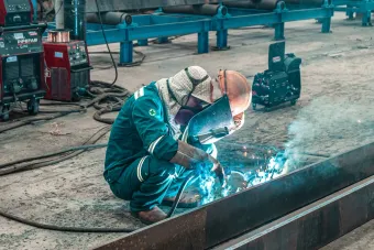 Welder in blue coveralls, kneeling, welding metal with sparks, industrial setting.