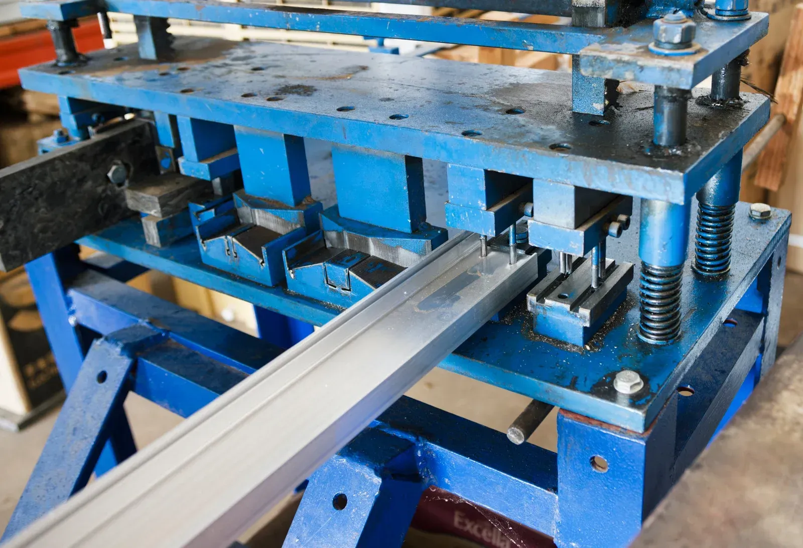 Blue metal forming machine shaping a silver aluminum extrusion in a workshop.
