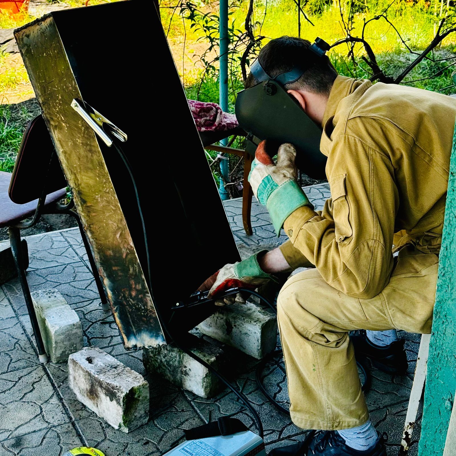 Person welds metal outdoors, wearing a welding mask and safety gear.
