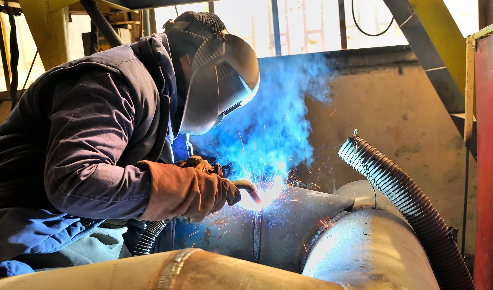 Welder in protective gear welding a large pipe, with bright blue arc and sparks.