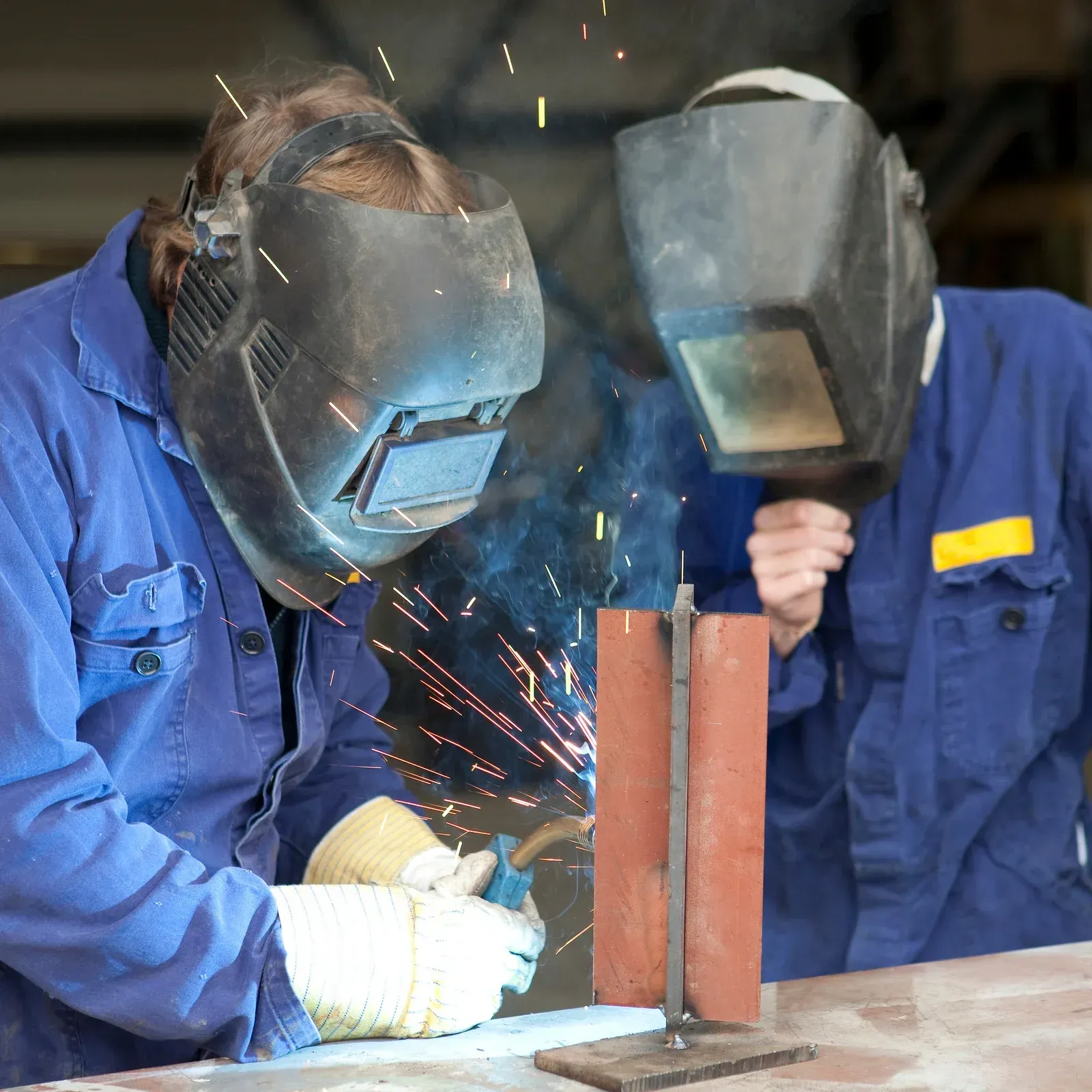 Two welders in blue coveralls and masks welding metal, sparks flying.
