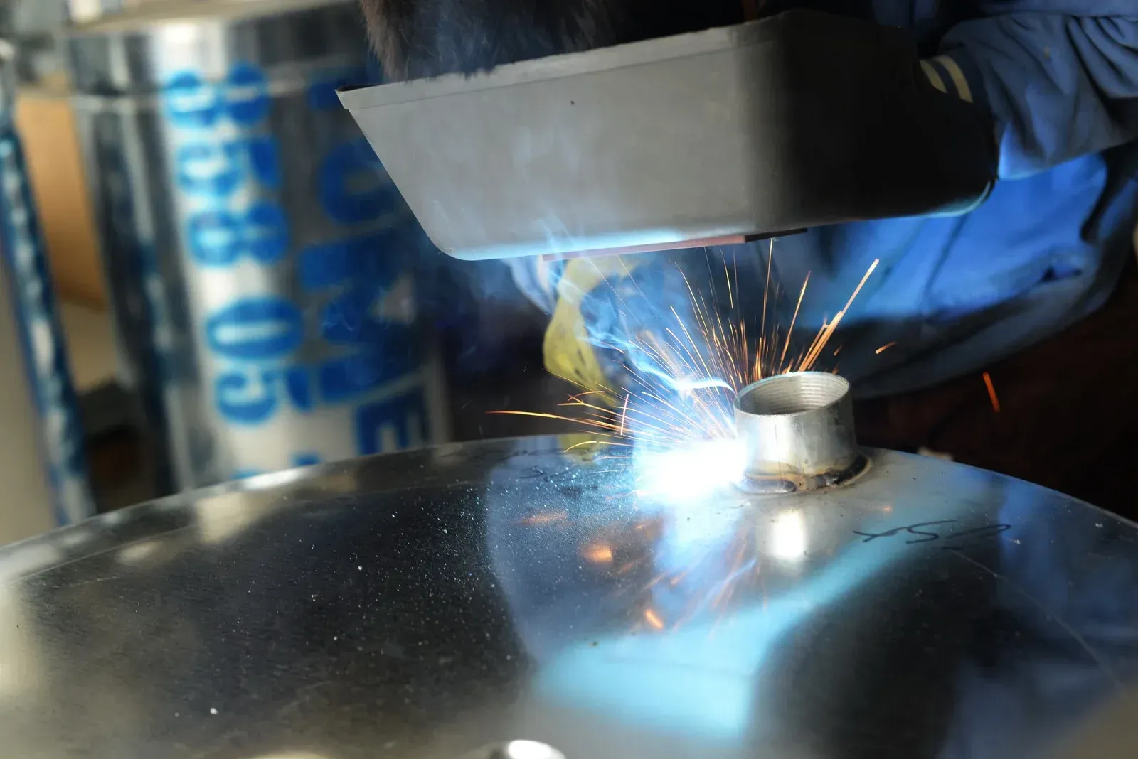 Welder in blue gloves and mask welding a metal piece, sparks flying.