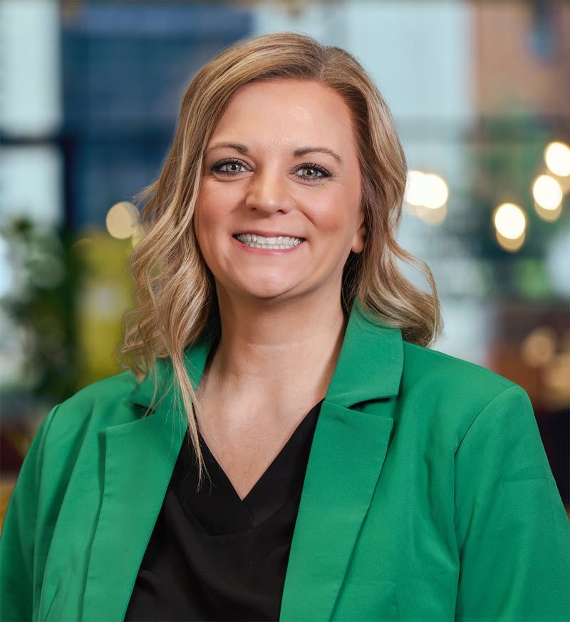 Woman in a green blazer smiles at the camera, blurred background.