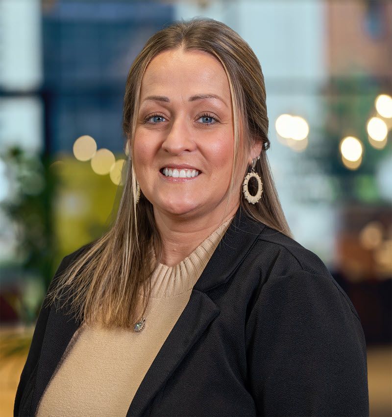 Woman smiling, wearing a black blazer, cream sweater, and dangling earrings. Blurred background.