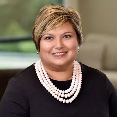 Woman in a green blazer smiles at the camera, blurred background.
