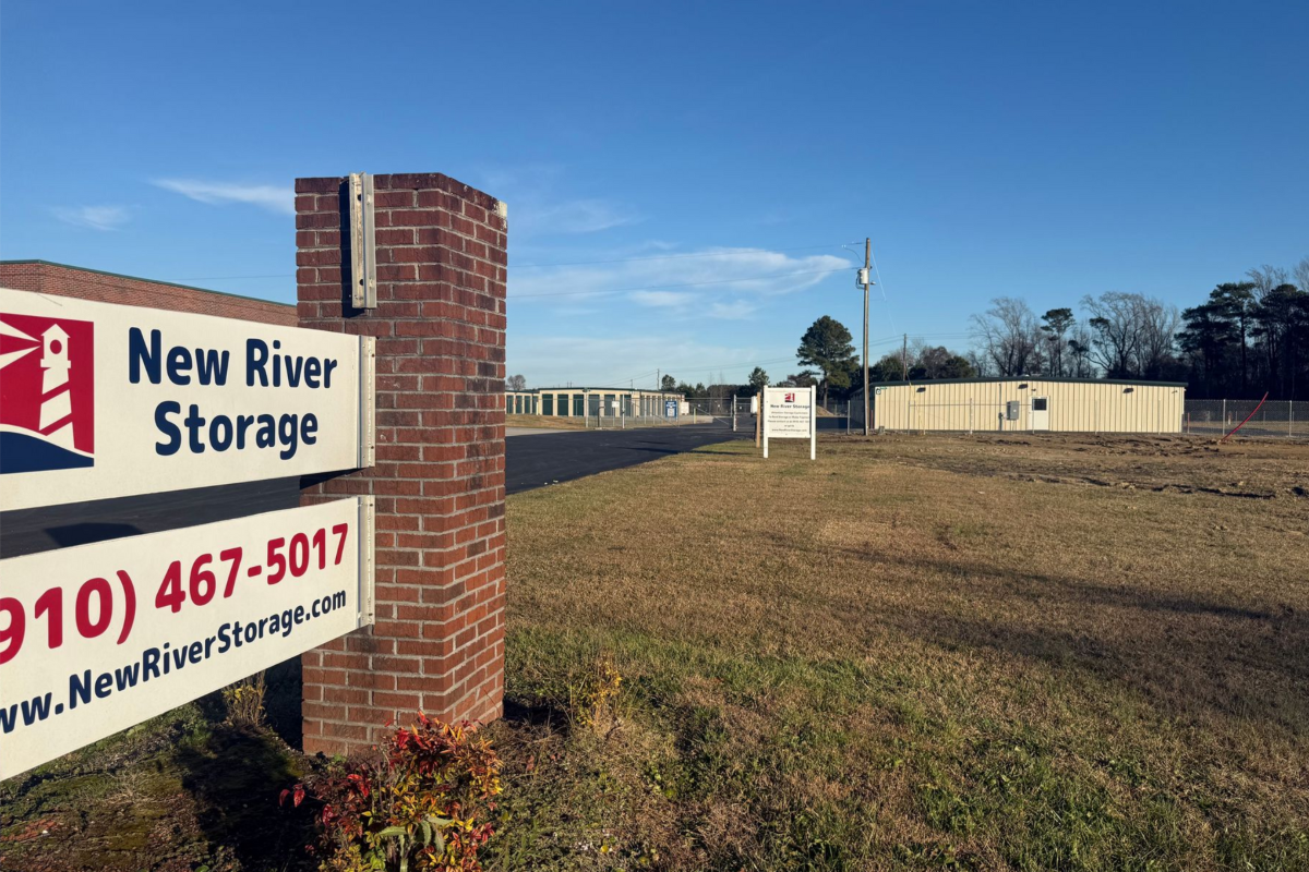 A brick sign for New River Storage with a phone number, overlooking a storage facility lot on a clear, sunny day.
