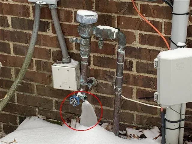 Ice buildup on exterior water valve, next to brick wall covered in snow.