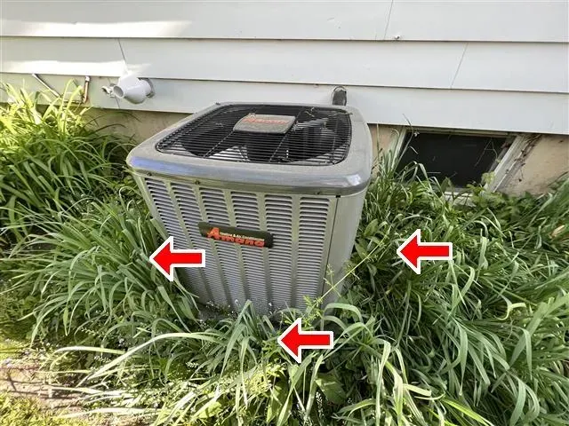 AC unit surrounded by tall grass next to a house, three red arrows pointing at the unit.