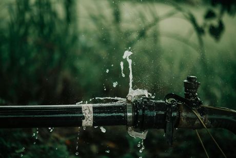 Water spurting from a broken black pipe, with a green, blurred background.