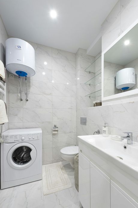 White bathroom with a washer, toilet, sink, and water heater on a marble-look tiled wall.