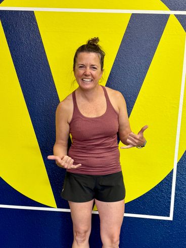 Woman in ribbed athletic tank top posing with a hand sign in front of Vitruvian Fitness logo