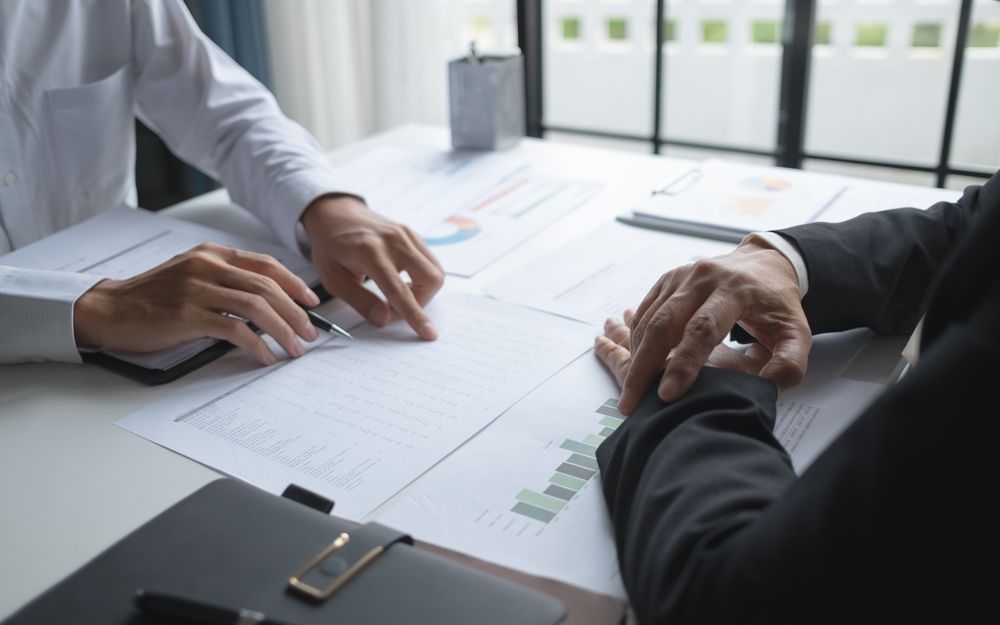 Two people in suits reviewing papers at a table, pointing at charts.